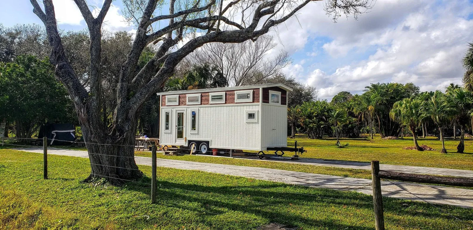 White tiny house on wheels beneath large trees.